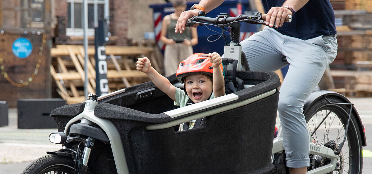Ein Lastenrad für den Kindertransport ist der Traum vieler junger Familien. Ein Kind in einem Lastenrad hebt jubelnd die Arme, das Lastenrad fährt eine erwachsene Person in Jeans. für den Kindertransport ist der Traum vieler junger Familien.