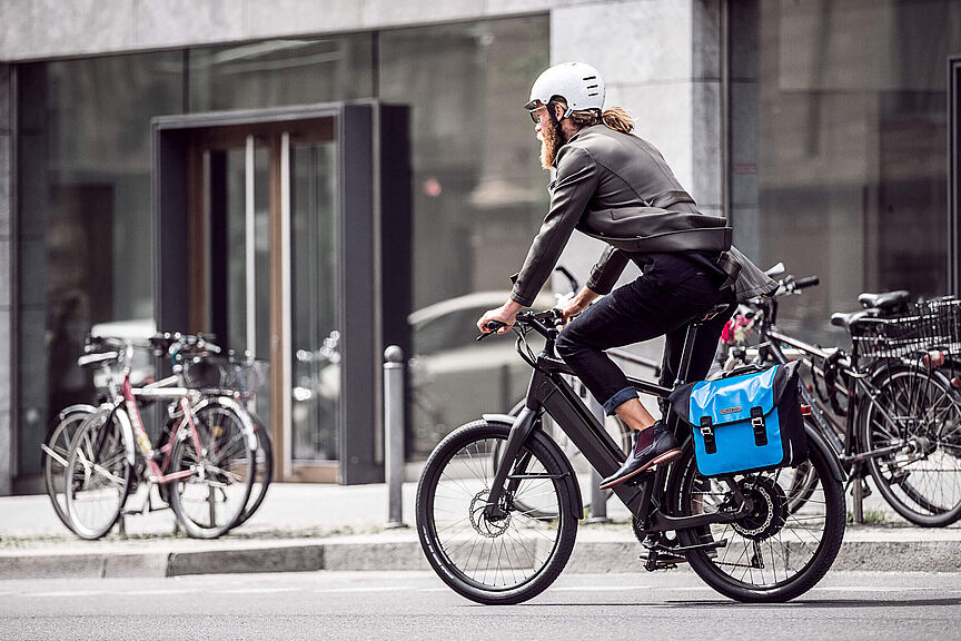 Berufspendler in der Stadt Ein Mann mit Bart und weißem Helm fährt auf einem modernen Elektrofahrrad durch eine städtische Straße. Er trägt Alltagskleidung (Jacke und Jeans) und hat eine leuchtend blaue Packtasche am Gepäckträger befestigt. Im Hintergrund sind Gebäude mit großen Glasfronten und parkende Fahrräder unscharf zu erkennen.
